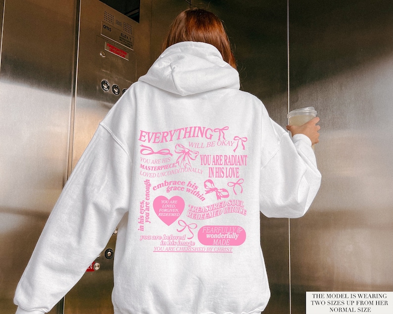 May include: White hoodie with pink text and bows. The text reads "Everything will be okay, you are his masterpiece, loved unconditionally, you are radiant in his love, you are enough in his eyes, embrace his grace within, you are loved, forgiven, redeemed, treasured soul, redeemed, you are holy in his hands, you are cherished by Christ, fearfully and wonderfully made".
