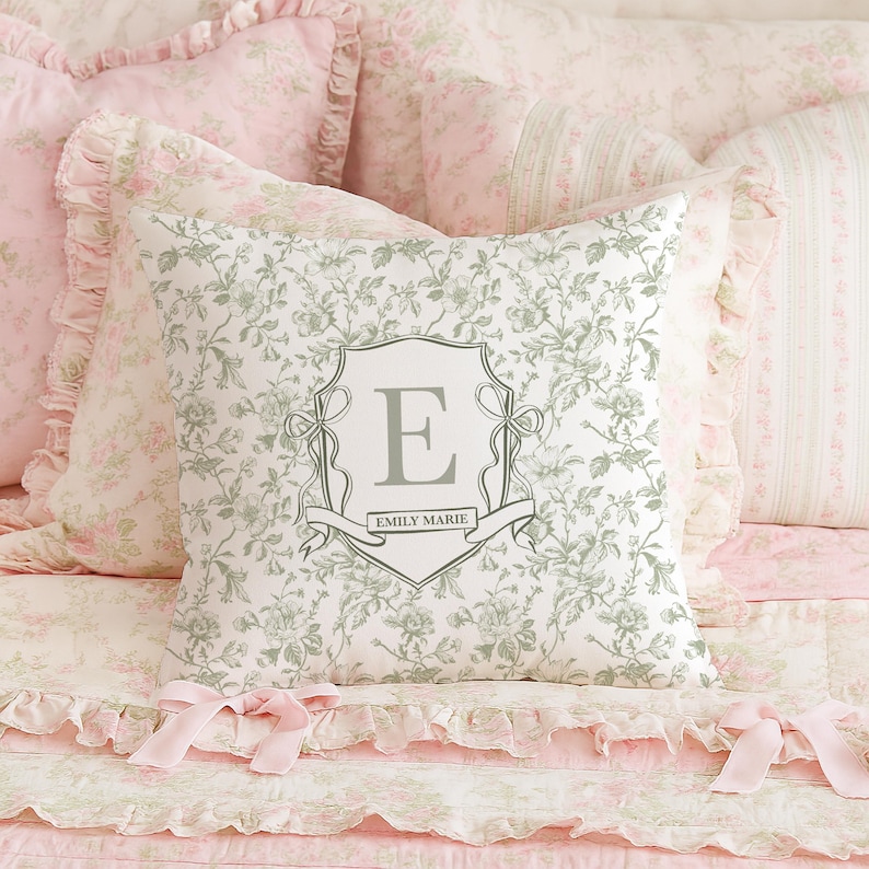 May include: A decorative pillow with a floral pattern and a gray monogrammed crest featuring the letter "E" and the name "Emily Marie". The pillow is surrounded by other pillows and bedding in shades of pink and white.