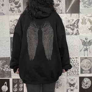 Pu&ograve; includere: Felpa con cappuccio nera oversize con una grafica di ali d'angelo argentate sul retro. Il modello indossa la felpa due taglie in pi&ugrave; rispetto alla sua taglia normale.