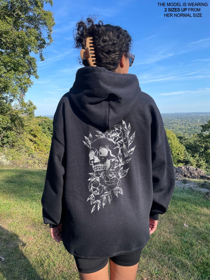 Puede incluir: Sudadera con capucha negra con un dise&ntilde;o de calavera blanca y flores en la espalda. La modelo lleva la sudadera dos tallas m&aacute;s grande que su talla normal.