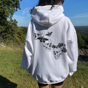 May include: White hooded sweatshirt with a black and white graphic print of three skulls with butterflies and flowers on the back.