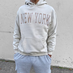 May include: A light beige hoodie with a large "NEW YORK" graphic in a brown and white color scheme. The model is wearing the hoodie in his normal size.