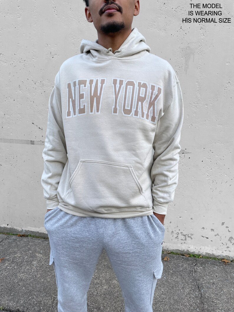 May include: A light beige hoodie with a large "NEW YORK" graphic in a brown and white color scheme. The model is wearing the hoodie in his normal size.