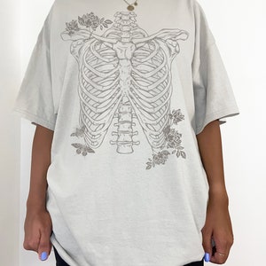 K&ouml;nnte beinhalten: Ein hellgraues Oversize-T-Shirt mit einem Grafikdruck eines Brustkorbs mit Blumen und Schmetterlingen. Das Model tr&auml;gt das Shirt zwei Gr&ouml;&szlig;en gr&ouml;&szlig;er als seine normale Gr&ouml;&szlig;e.