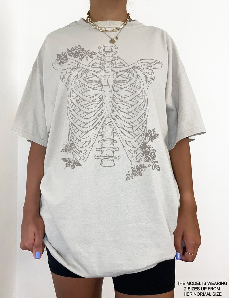 K&ouml;nnte beinhalten: Ein hellgraues Oversize-T-Shirt mit einem Grafikdruck eines Brustkorbs mit Blumen und Schmetterlingen. Das Model tr&auml;gt das Shirt zwei Gr&ouml;&szlig;en gr&ouml;&szlig;er als seine normale Gr&ouml;&szlig;e.