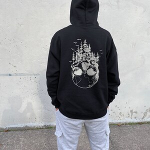 May include: Black hooded sweatshirt with a white graphic print of a globe with trees and birds on it. The model is wearing the sweatshirt with grey sweatpants.