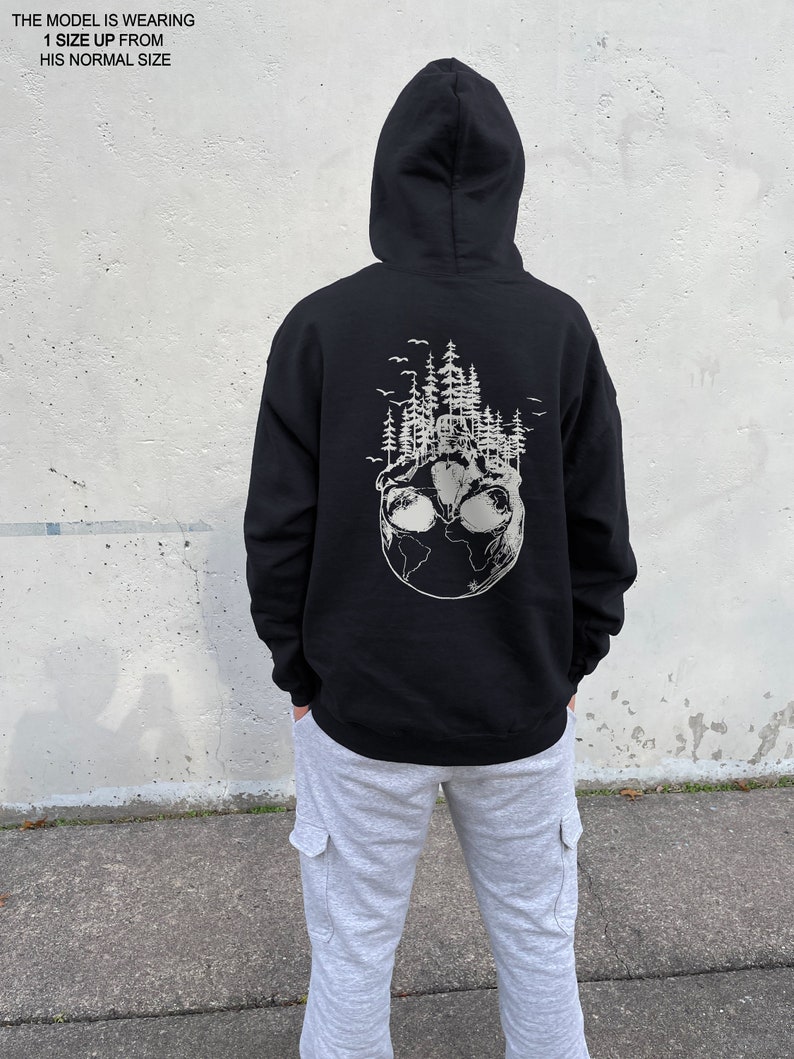 May include: Black hooded sweatshirt with a white graphic print of a globe with trees and birds on it. The model is wearing the sweatshirt with grey sweatpants.
