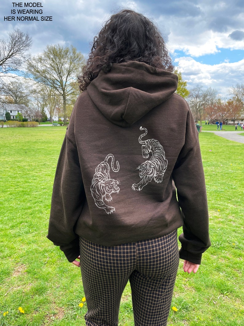 May include: A brown hoodie with a white graphic of two tigers facing each other on the back. The model is wearing the hoodie with brown and black checked trousers.