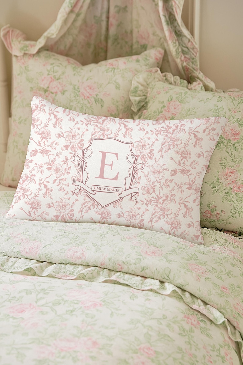 May include: A bed set with a floral pattern in pink and green. The focal point is a rectangular pillow with a pink floral design and a shield with the letter "E" and the name "EMILY MARIE".