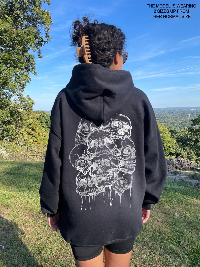 May include: Black hooded sweatshirt with a graphic print of multiple white skulls dripping with white paint. The model is wearing the sweatshirt two sizes up from her normal size.