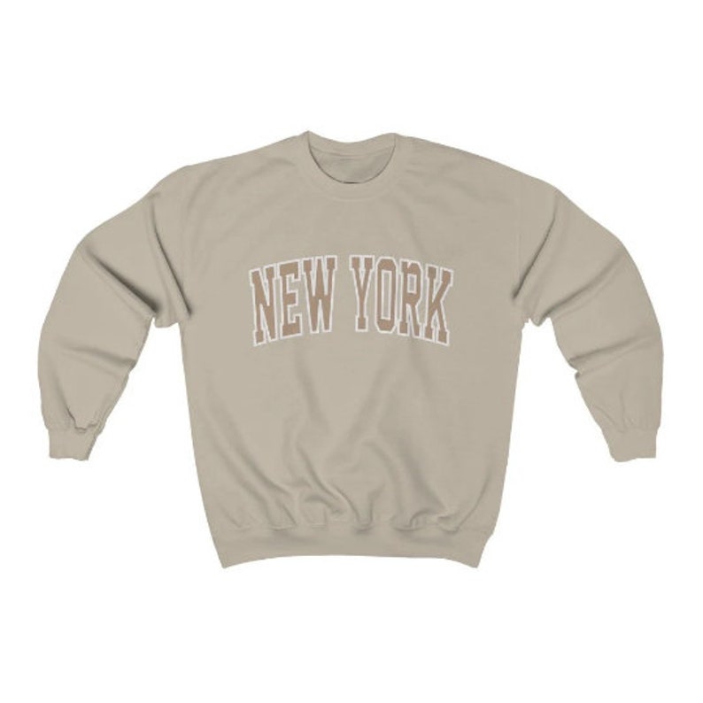 May include: A beige sweatshirt with the text "NEW YORK" in brown letters with a white outline.