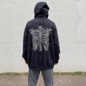 May include: Black hooded sweatshirt with a white skeleton print on the back. The model is wearing the sweatshirt one size up from their normal size.