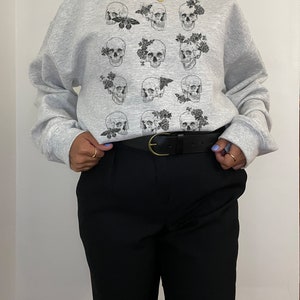 K&ouml;nnte beinhalten: Grauer Pullover mit einem Druck von mehreren schwarzen und wei&szlig;en Totenk&ouml;pfen mit Blumen und Schmetterlingen.