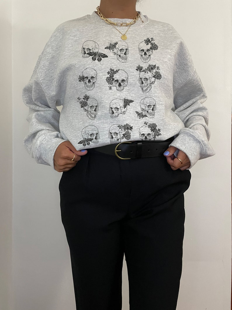 K&ouml;nnte beinhalten: Grauer Pullover mit einem Druck von mehreren schwarzen und wei&szlig;en Totenk&ouml;pfen mit Blumen und Schmetterlingen.