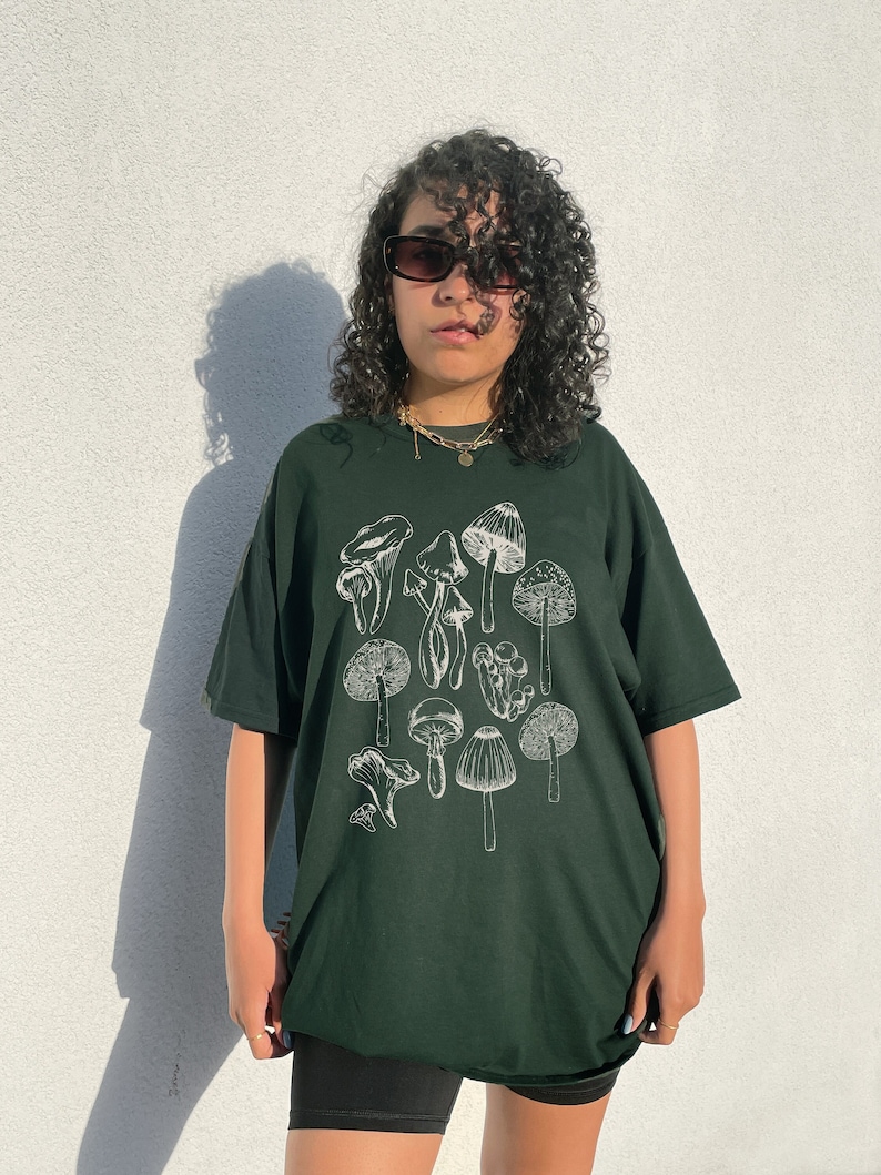 May include: A dark green t-shirt with a white graphic of various mushrooms. The shirt has a relaxed fit and a crew neck.