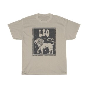 K&ouml;nnte beinhalten: Ein beiges T-Shirt mit einer Grafik im Vintage-Stil eines L&ouml;wen und dem Wort "LEO" in einer Retro-Schrift.