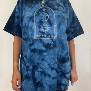 May include: A blue tie-dye t-shirt with a white graphic of a sun, moon, and stars in an archway. The text "XXIV" and "VENI. VIDI. AMAVI." are printed on the shirt.