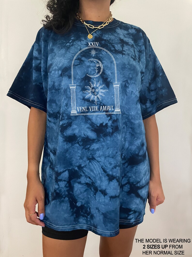 May include: A blue tie-dye t-shirt with a white graphic of a sun, moon, and stars in an archway. The text "XXIV" and "VENI. VIDI. AMAVI." are printed on the shirt.