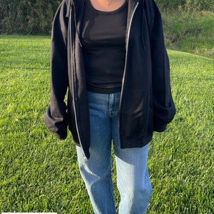 May include: A black zip-up hoodie with a hood and a zipper. The model is wearing the hoodie with the zipper open. The model is wearing blue jeans and a black tank top underneath the hoodie. The text "THE MODEL IS WEARING 2 SIZES UP FROM HER NORMAL SIZE" is at the bottom of the image.