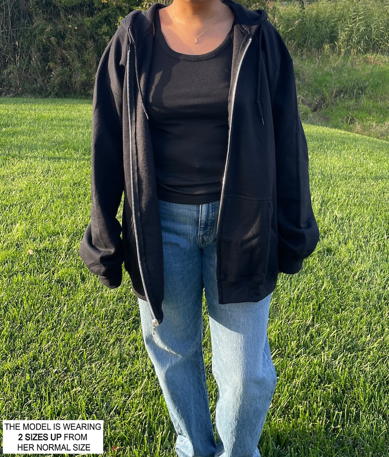 May include: A black zip-up hoodie with a hood and a zipper. The model is wearing the hoodie with the zipper open. The model is wearing blue jeans and a black tank top underneath the hoodie. The text "THE MODEL IS WEARING 2 SIZES UP FROM HER NORMAL SIZE" is at the bottom of the image.
