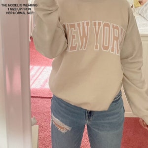 May include: A beige sweatshirt with the text "NEW YORK" printed in white with a pink outline. The model is wearing the sweatshirt with blue jeans.
