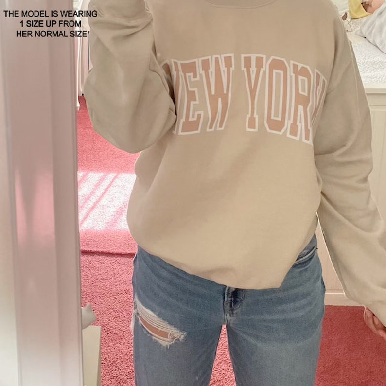 May include: A beige sweatshirt with the text "NEW YORK" printed in white with a pink outline. The model is wearing the sweatshirt with blue jeans.