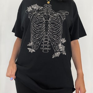 K&ouml;nnte beinhalten: Schwarzes Oversize-T-Shirt mit einem wei&szlig;en Aufdruck eines Brustkorbs mit Blumen und Schmetterlingen.