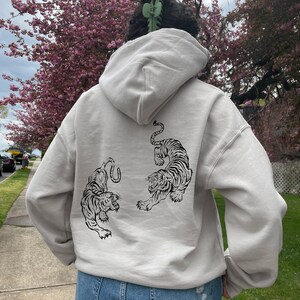 May include: A light gray hooded sweatshirt with a black graphic of two tigers on the back. The model is wearing the sweatshirt with the hood up.