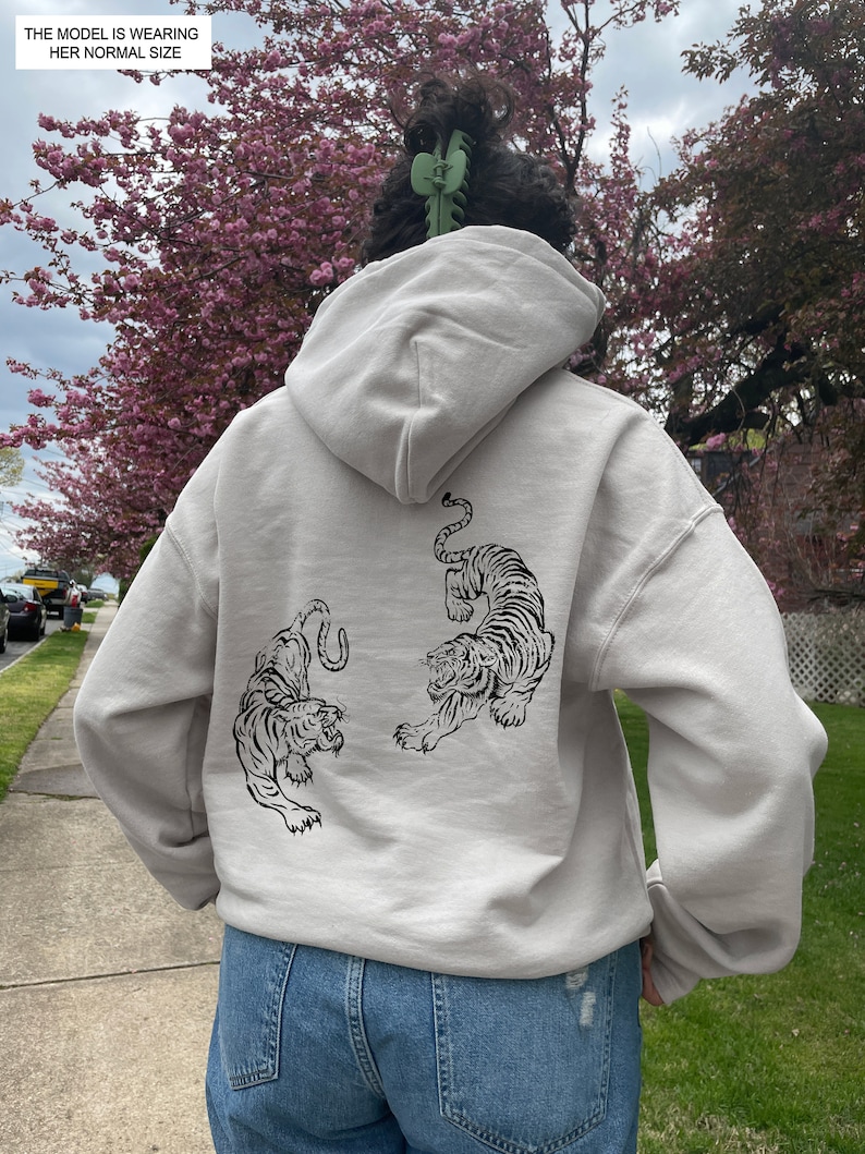 May include: A light gray hooded sweatshirt with a black graphic of two tigers on the back. The model is wearing the sweatshirt with the hood up.