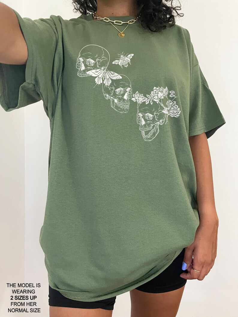 Puede incluir: Una camiseta verde de gran tama&ntilde;o con un estampado gr&aacute;fico blanco de tres calaveras con flores y una mariposa. La modelo lleva la camiseta dos tallas m&aacute;s grande que su talla normal.