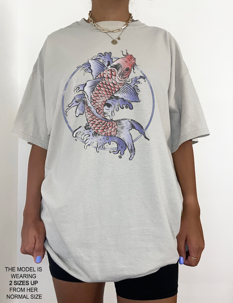 May include: A light gray oversized t-shirt with a graphic print of a red and blue koi fish swimming in a circle of water. The model is wearing the shirt two sizes up from her normal size. "THE MODEL IS WEARING 2 SIZES UP FROM HER NORMAL SIZE" is printed on the bottom of the image.