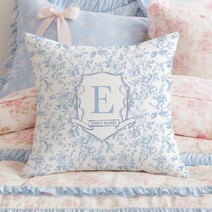 May include: A square decorative pillow with a floral pattern in white and light blue. The pillow features a central crest design with the letter "E" and the name "EMILY MARIE" in light blue. Other pillows and bedding are visible in the background.