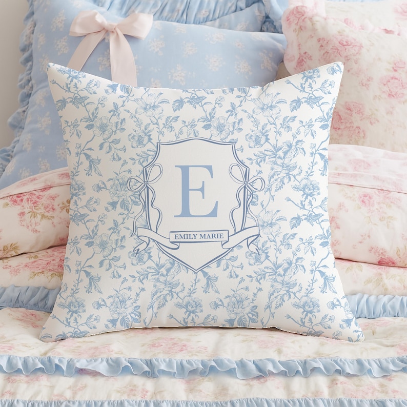 May include: A square decorative pillow with a floral pattern in white and light blue. The pillow features a central crest design with the letter "E" and the name "EMILY MARIE" in light blue. Other pillows and bedding are visible in the background.