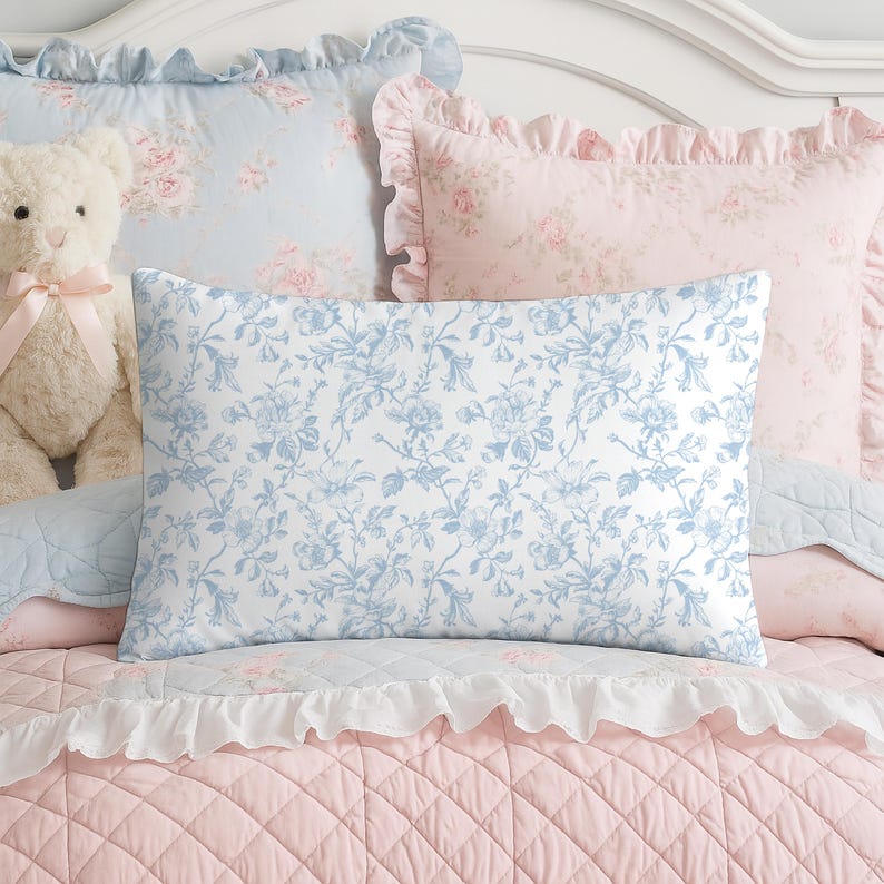 May include: A collection of decorative pillows and bedding in soft pastel colors. The rectangular pillow in the foreground features a blue floral pattern on a white background. Other pillows have ruffled edges and floral designs in pink and blue. A teddy bear is visible.