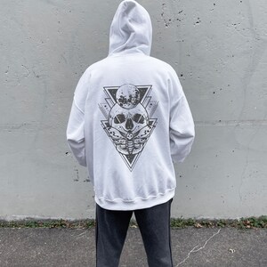 May include: White hooded sweatshirt with a black and white graphic print of a skull with a moth and a crescent moon inside a triangle on the back.