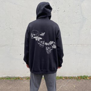 May include: Black hooded sweatshirt with a graphic print of three skulls with flowers and butterflies. The model is wearing the sweatshirt one size up from their normal size.