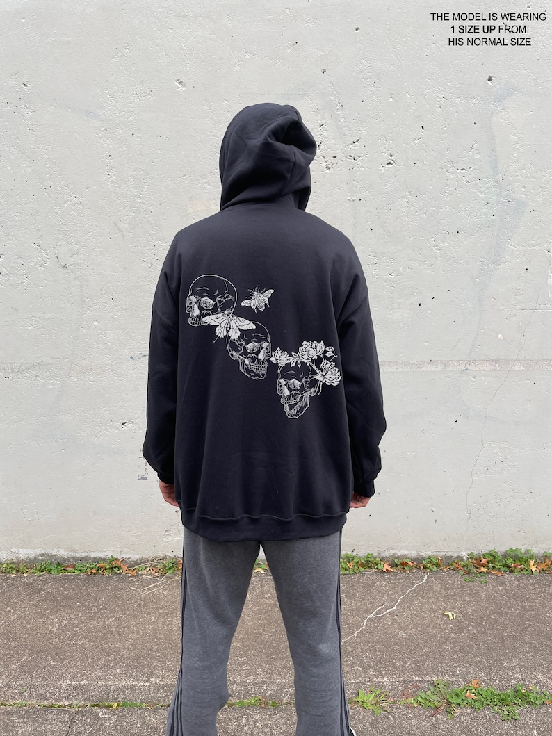 May include: Black hooded sweatshirt with a graphic print of three skulls with flowers and butterflies. The model is wearing the sweatshirt one size up from their normal size.