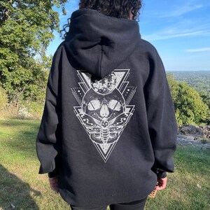 May include: Black hooded sweatshirt with a white graphic print of a skull with a moth and geometric shapes. The model is wearing the sweatshirt two sizes up from her normal size.