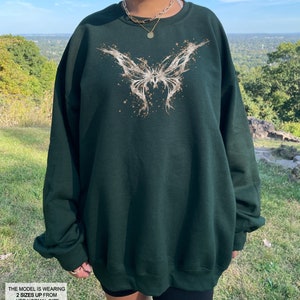 Pode incluir: Uma camisola verde escura com um gr&aacute;fico de borboleta branca. A borboleta &eacute; estilizada com um efeito de pintura salpicada. A modelo est&aacute; usando um tamanho dois tamanhos acima do seu tamanho normal.