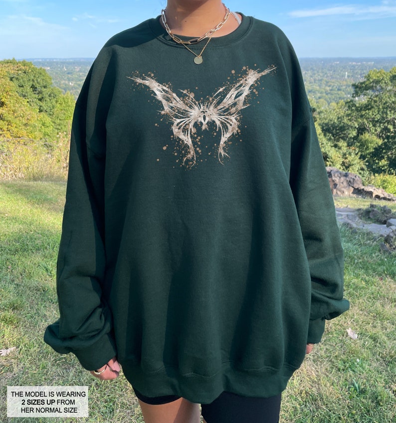 Pode incluir: Uma camisola verde escura com um gr&aacute;fico de borboleta branca. A borboleta &eacute; estilizada com um efeito de pintura salpicada. A modelo est&aacute; usando um tamanho dois tamanhos acima do seu tamanho normal.