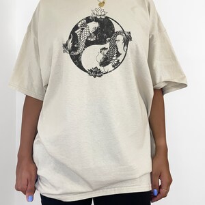 May include: A beige oversized t-shirt with a black yin yang symbol featuring two fish and lotus flowers. The model is wearing the shirt two sizes up from her normal size.