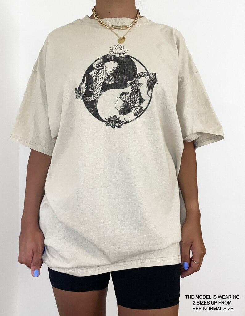 May include: A beige oversized t-shirt with a black yin yang symbol featuring two fish and lotus flowers. The model is wearing the shirt two sizes up from her normal size.