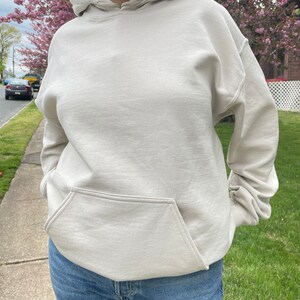May include: A light beige hoodie with a kangaroo pocket. The hoodie has a drawstring hood and long sleeves.