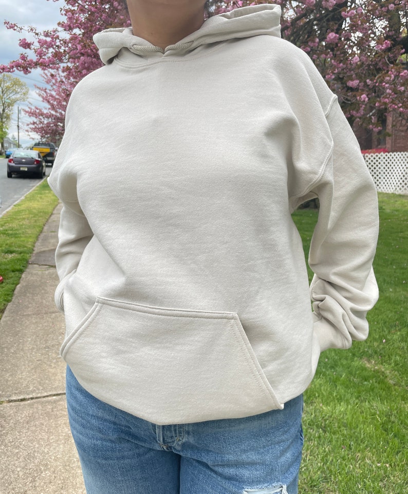 May include: A light beige hoodie with a kangaroo pocket. The hoodie has a drawstring hood and long sleeves.