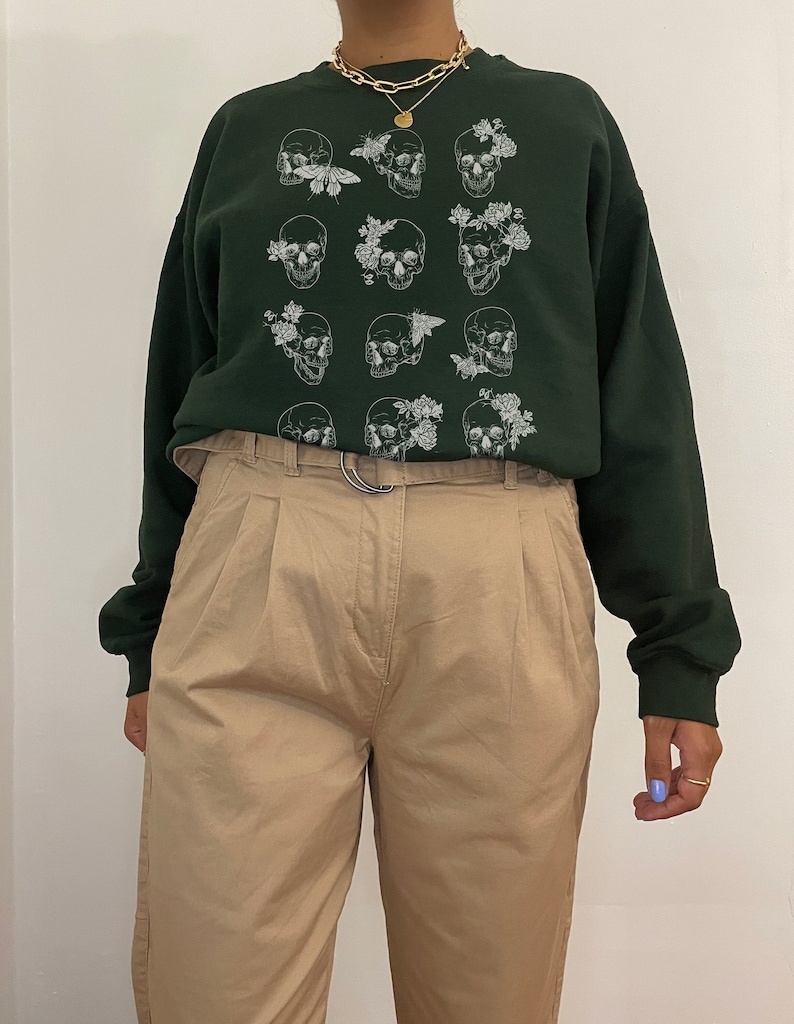 K&ouml;nnte beinhalten: Ein dunkelgr&uuml;ner Pullover mit einem wei&szlig;en Druck von mehreren Sch&auml;deln mit floralen Akzenten und Schmetterlingen.