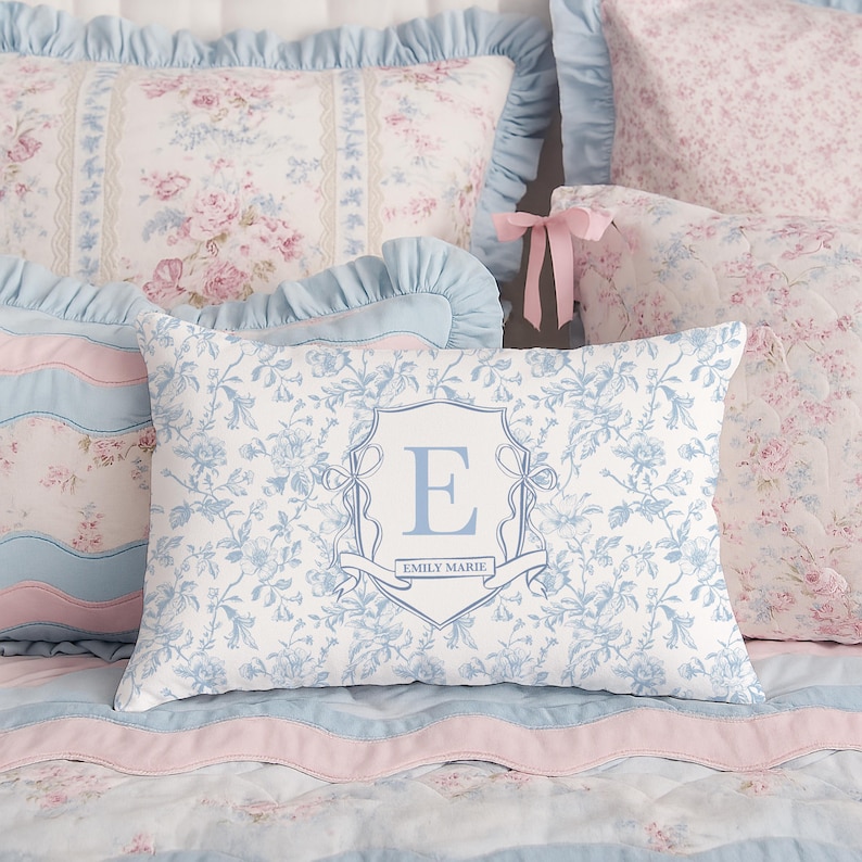 May include: A collection of decorative pillows in shades of blue, pink, and white. The pillows feature floral patterns and a monogrammed pillow with the letter "E" and the name "Emily Marie". The bedding has a romantic, vintage aesthetic.