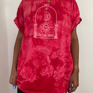 May include: Red tie-dye t-shirt with a white graphic of an archway with a sun and moon inside. The text "XXIV" and "VENI. VIDI. AMAVI." are printed on the shirt.