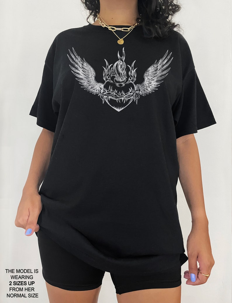 May include: Black oversized t-shirt featuring a graphic print of a flaming heart with wings and barbed wire. The model is wearing the shirt two sizes up from her usual size.