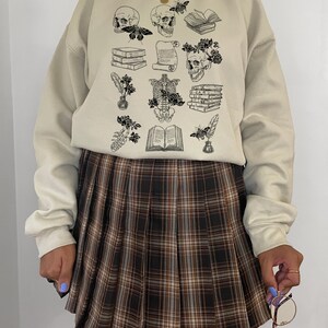 May include: A beige sweatshirt with a black and white graphic print of skulls, books, flowers, and other gothic-inspired imagery. The sweatshirt is paired with a brown plaid pleated skirt.