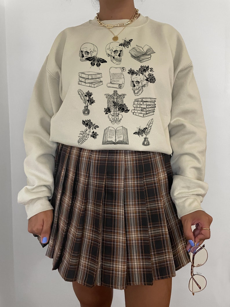 May include: A beige sweatshirt with a black and white graphic print of skulls, books, flowers, and other gothic-inspired imagery. The sweatshirt is paired with a brown plaid pleated skirt.